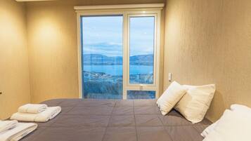 A bedroom with a bed, pillows, and a view of a body of water and landscape through a glass door.
