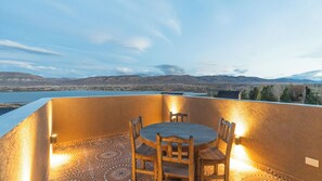 Una terraza con mesa y sillas, con vista a un cuerpo de agua y montañas a lo lejos.