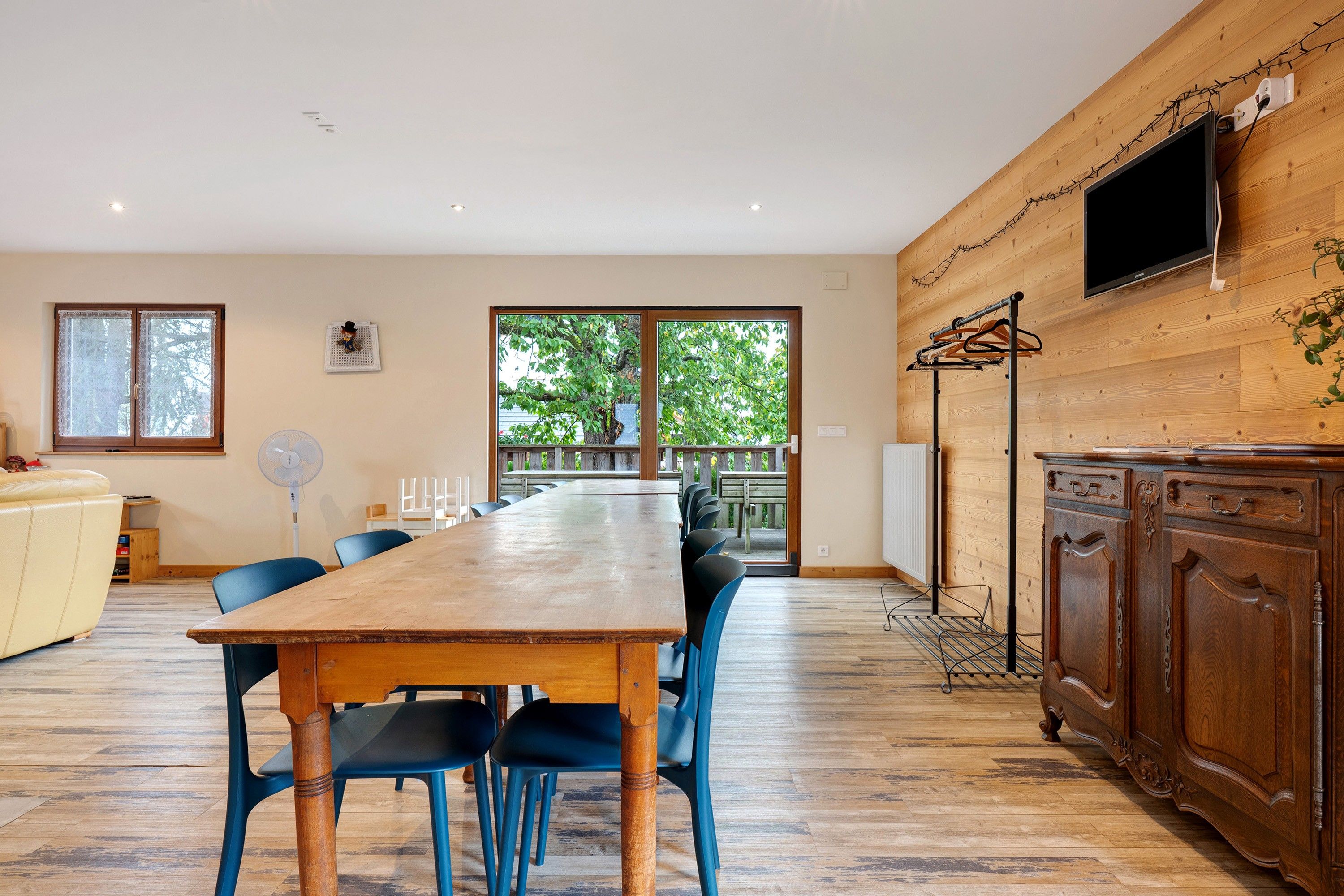 Un vaste espace de salle à manger avec une table en bois et des chaises bleues, un meuble en bois abritant un téléviseur, et une vue sur l’extérieur grâce à de grandes fenêtres.