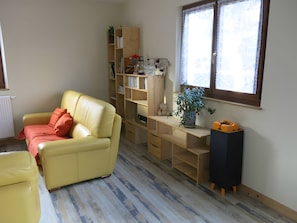 Ein Wohnzimmer mit einem gelben Sofa, Holzregalen, einem Blumentopf und einem Fenster mit Spitzenvorhängen.