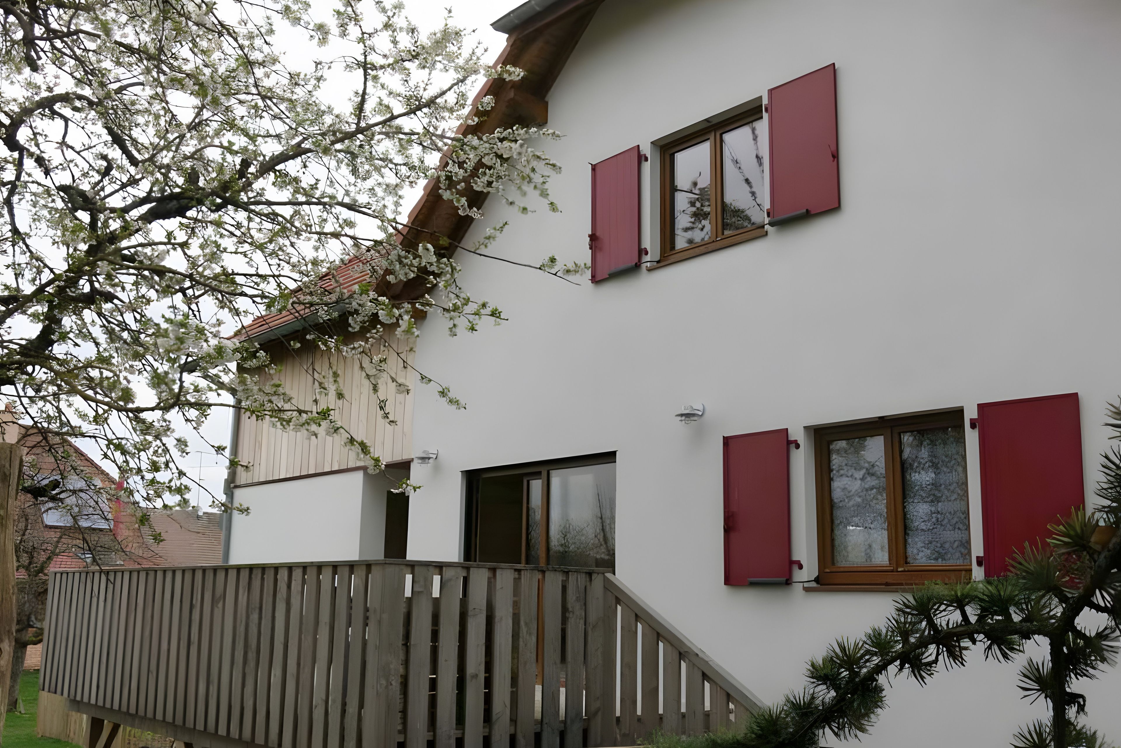 Un bâtiment à deux étages avec des volets rouges, un balcon en bois et un arbre aux fleurs blanches.