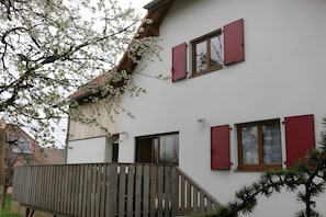 Ein zweistöckiges Gebäude mit roten Fensterläden, einem hölzernen Balkon und einem Baum mit weißen Blüten.