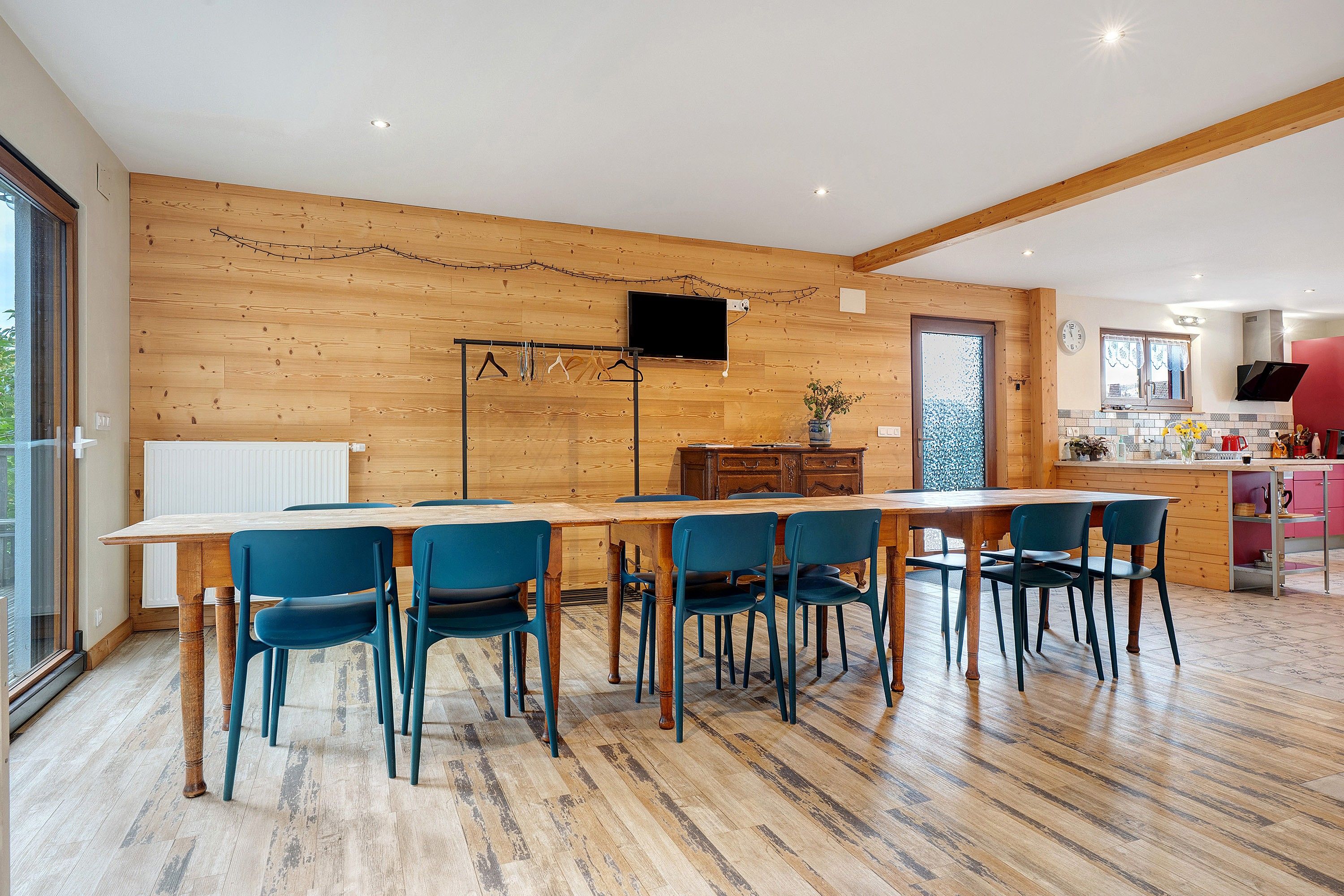 A dining area with a wooden table and chairs, a television mounted on the wall, and a kitchen in the background.