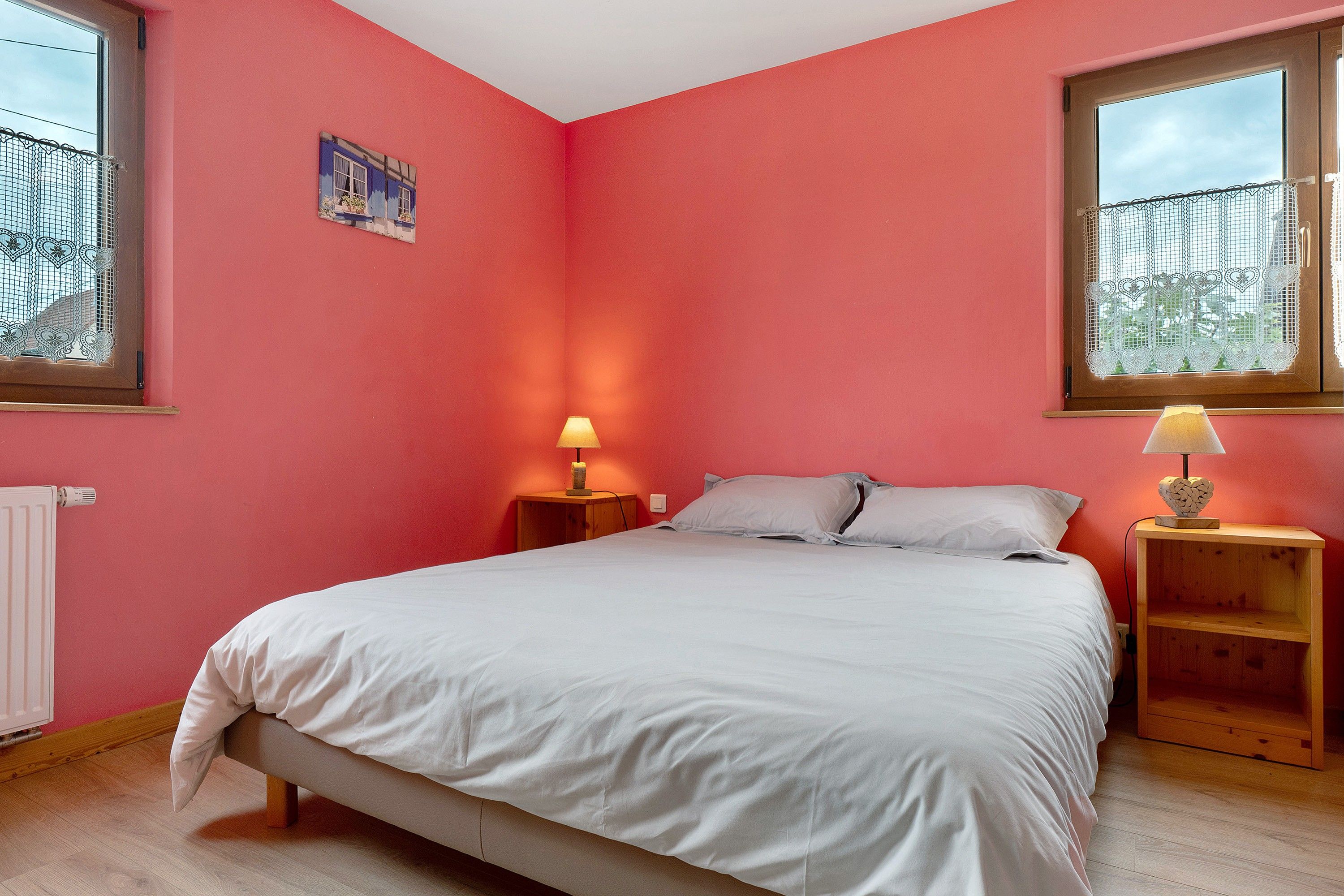 Un dormitorio con una cama, dos mesitas de noche, un radiador y una ventana con cortinas de encaje.