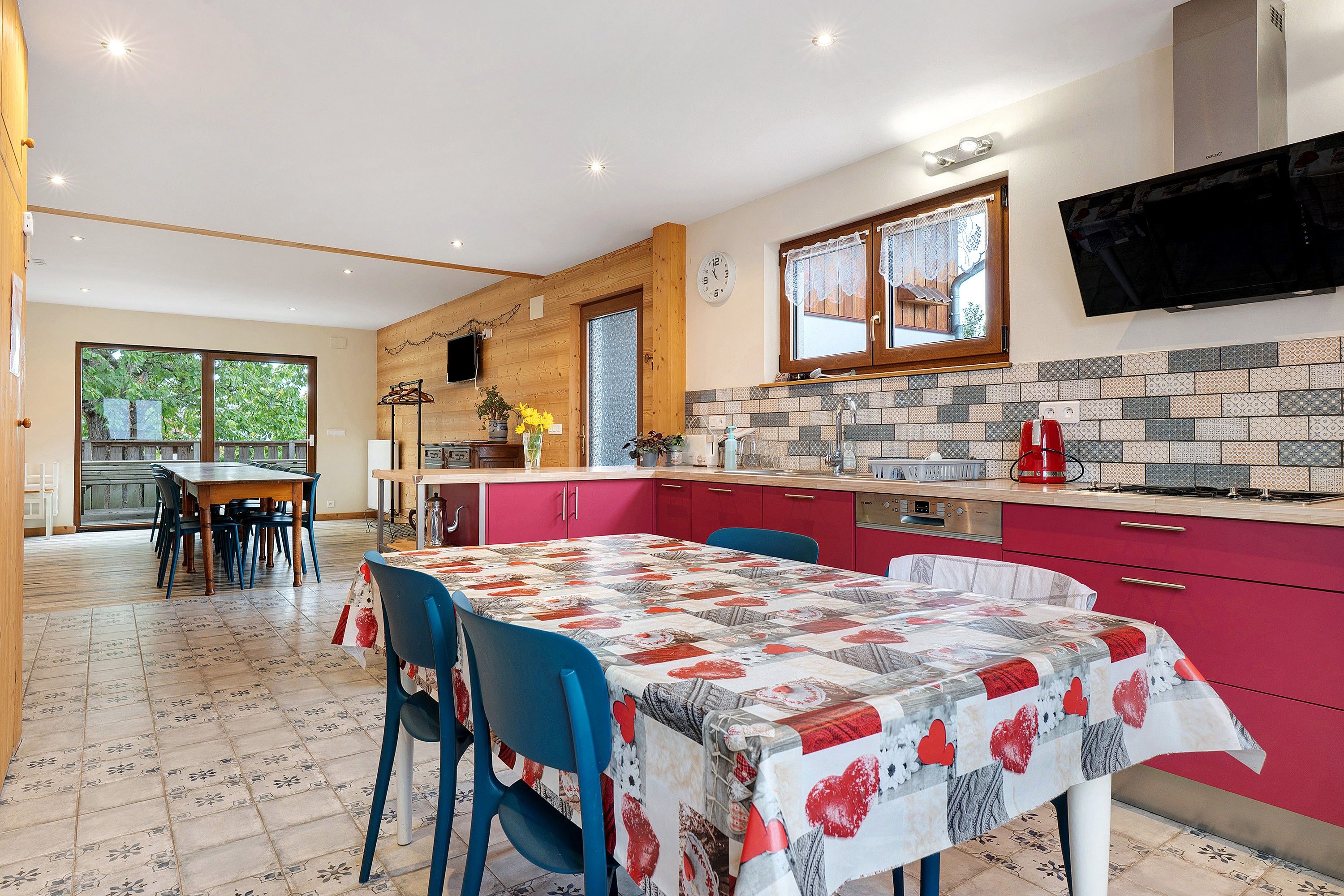 A modern kitchen with red cabinets, a tiled backsplash, and a dining area with a quilted tablecloth.
