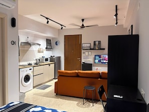 Un espai habitable reduït amb petita cuina, sofà, televisió i ventilador de sostre.