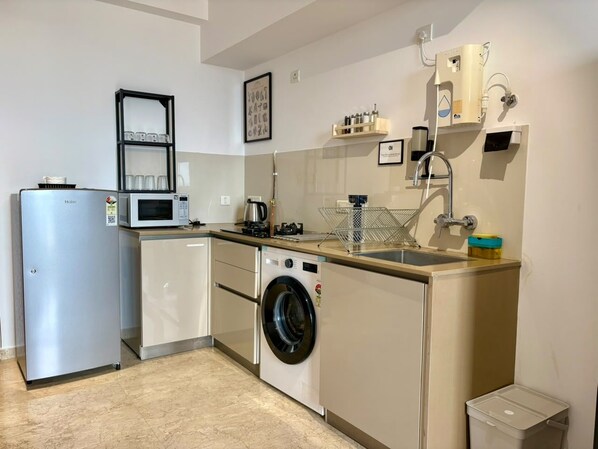 A compact kitchen with a refrigerator, microwave, washing machine, sink, and a small trash bin.
