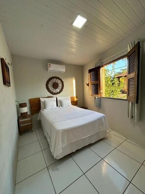 A bedroom with a bed, a bedside table, a wall-mounted air conditioner, and a window with shutters. - Vagner's house (Grajaú)