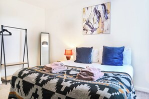 A hotel room with a bed, a bedside table with a lamp, a wall-mounted artwork, and a clothing rack. - Cosy Flat in Prestwich, Sleeps 4, Spacious, Work, Getaway (Prestwich)