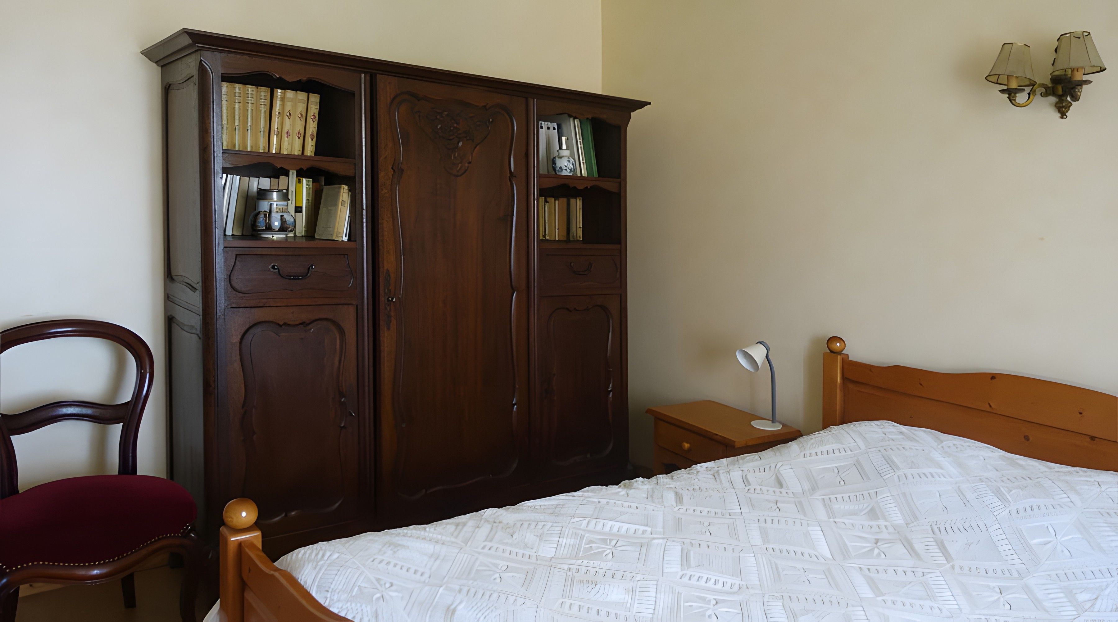 Une chambre avec une armoire en bois, un lit simple recouvert d’une couvre-lit à motifs, une table de chevet avec une lampe et une chaise.