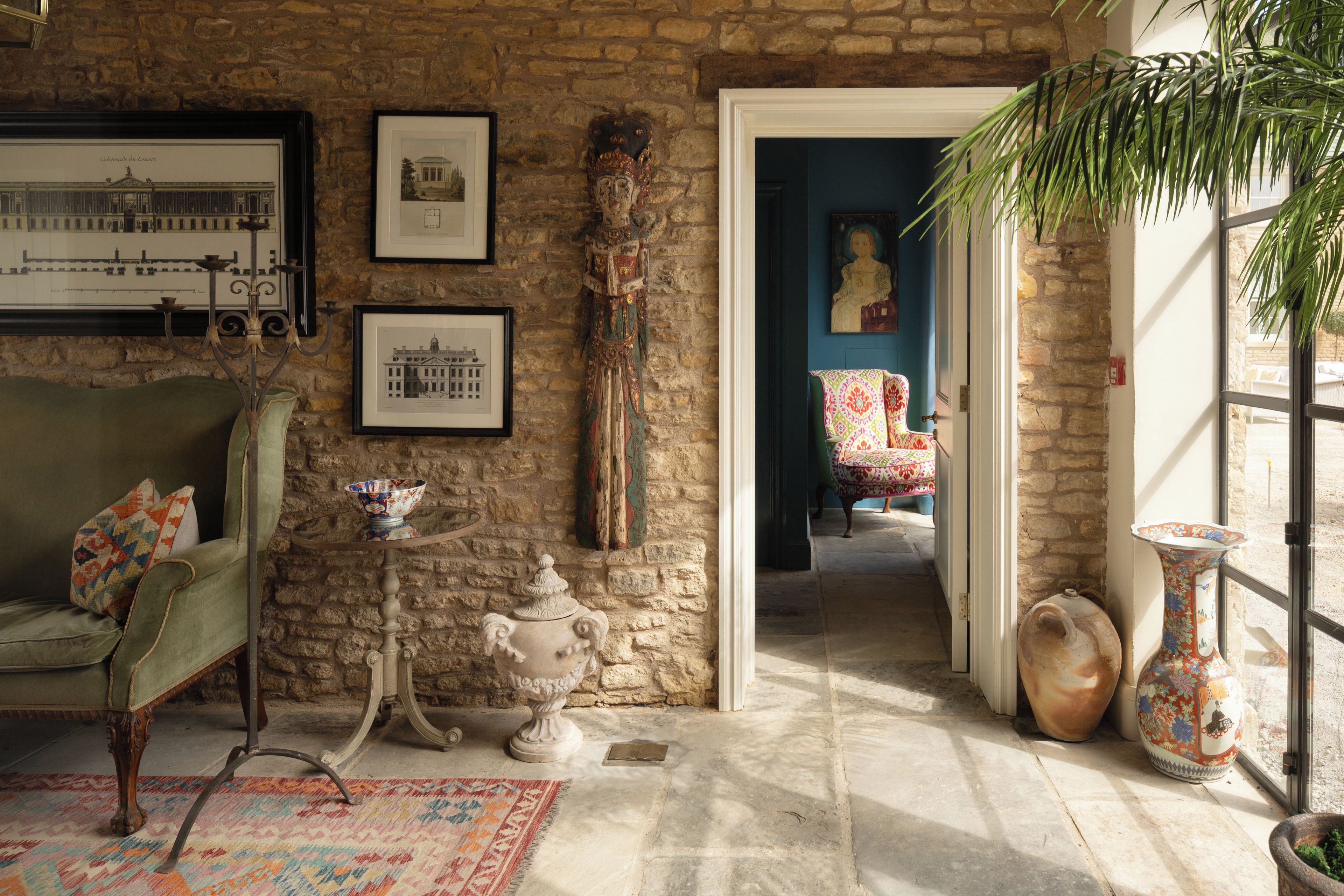 Una habitación con pared de piedra, un sillón verde, una mesita con un cuenco, y una puerta que da a otra habitación.