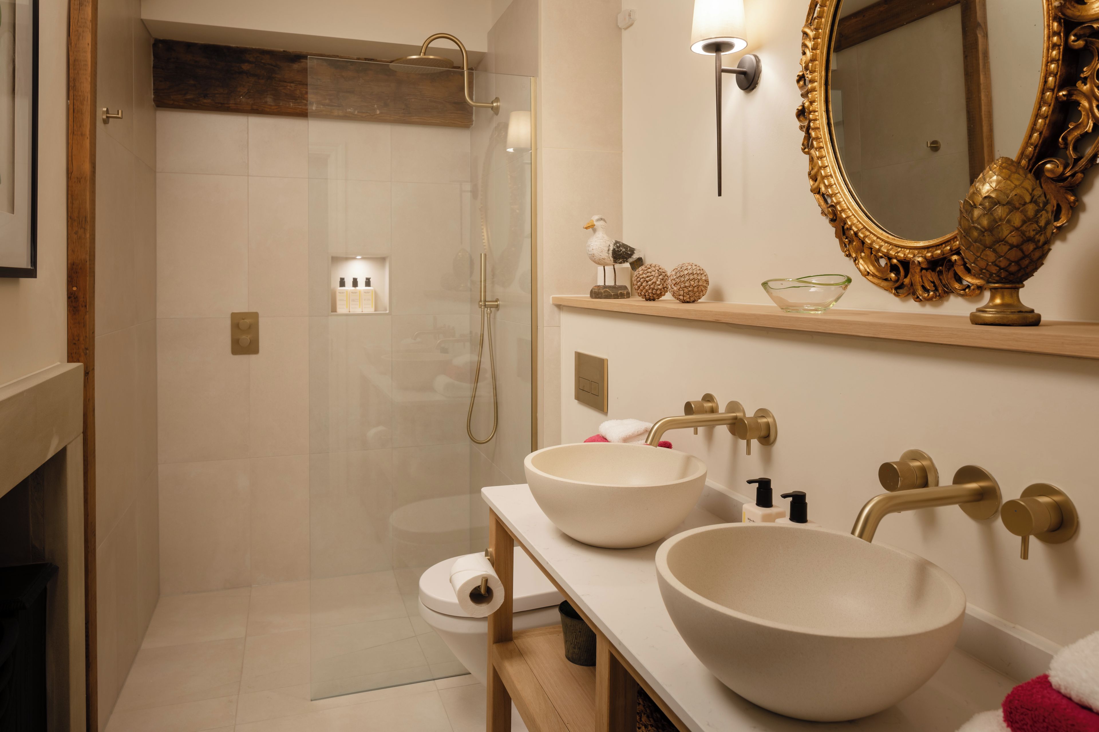 Un baño moderno con ducha de vidrio, dos lavamanos, un espejo con marco decorativo y un estante con adornos.