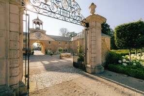 A gated entrance with a clock tower, cobblestone path, and a garden with white flowers.