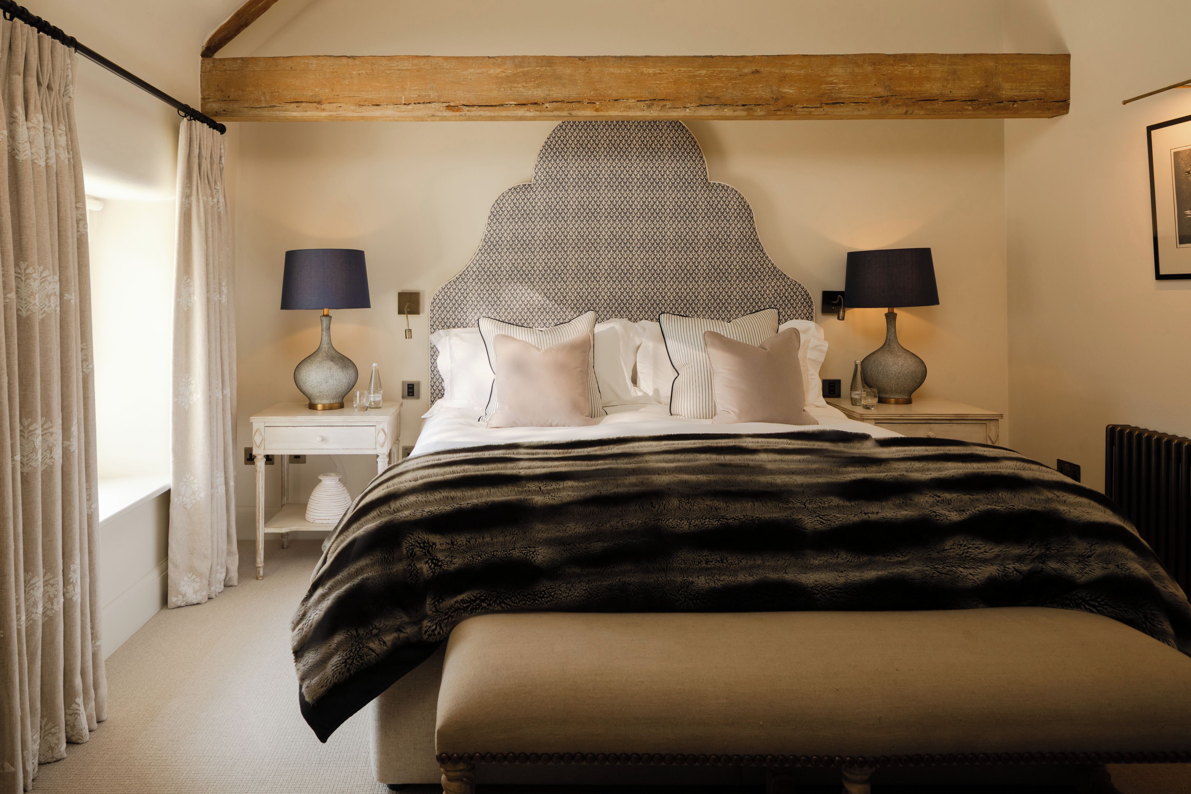 A bedroom with a large bed, two bedside tables with lamps, a window with curtains, and a radiator.