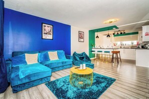 A modern living room with a blue sofa, a yellow coffee table, and a kitchen area visible in the background.