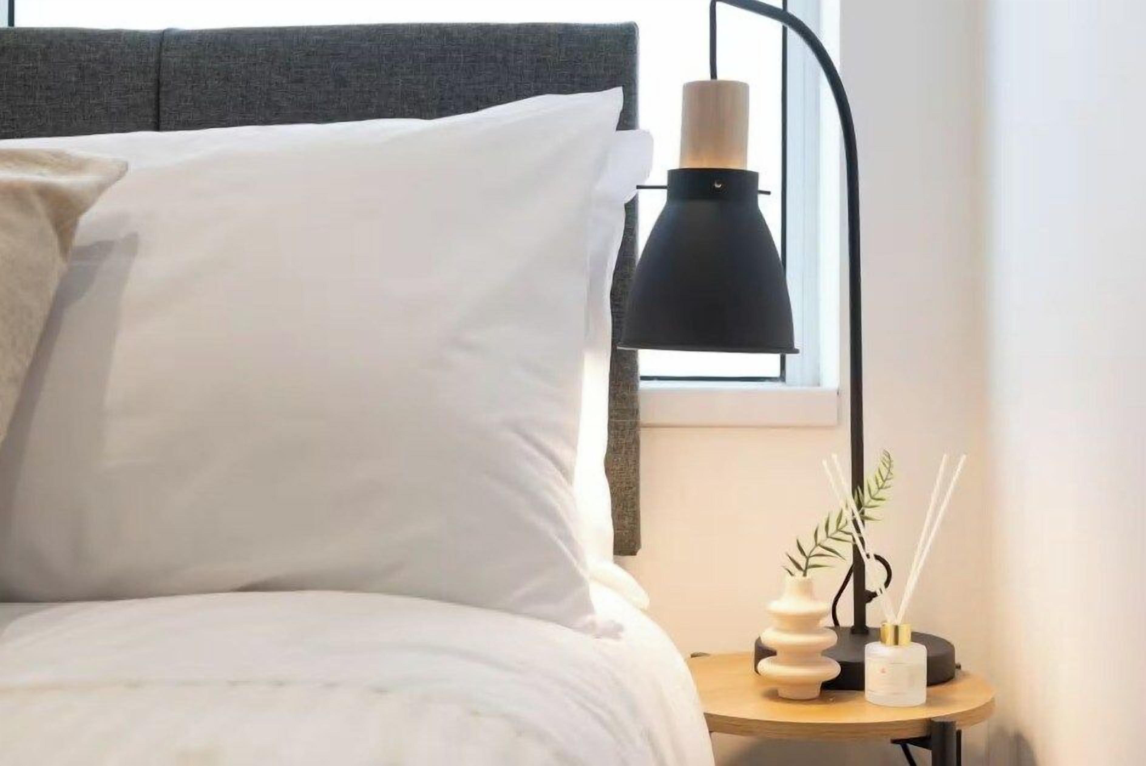 Una cama con ropa de cama blanca, una lámpara negra y una mesita con objetos decorativos.