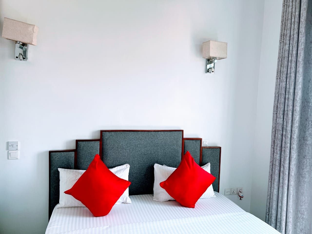 A hotel room with a bed, two red pillows, a grey headboard, and a grey curtain.