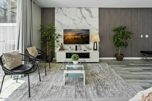 Ein modernes Wohnzimmer mit Flachbildfernseher, einer Couch, einem Couchtisch und zwei Zimmerpflanzen.