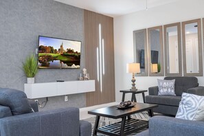 A modern living room with a flat-screen TV mounted on the wall, a sofa, a coffee table, and a lamp. - 4BR Townhome Near Disney Pool & Mickey Room (Kissimmee)