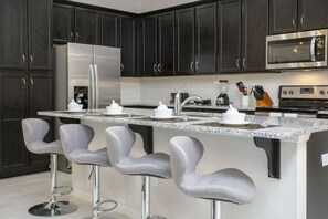 A modern kitchen with dark cabinets, stainless steel appliances, a granite countertop, and bar stools. - 5BR Solara Home Private Pool & Themed Rooms (Kissimmee)