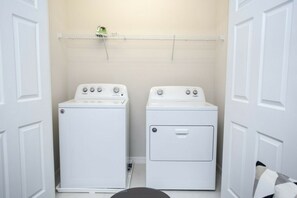 A laundry room with two white washers and dryers, a metal shelf above the dryers, and a white door on the left. - 5BR Solara Home Private Pool & Themed Rooms (Kissimmee)