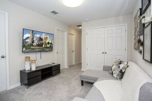 A living room with a flat-screen TV, a grey sofa, a black entertainment unit, and a hallway leading to a door. - 5BR Solara Home Private Pool & Themed Rooms (Kissimmee)