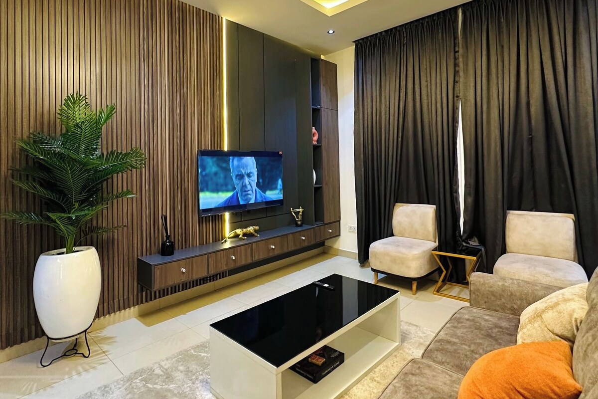 Uma sala de estar moderna com televisão de tela plana, uma mesa de centro, sofás e uma luminária decorativa no teto.