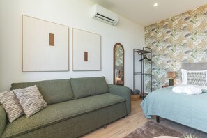 A bedroom with a green sofa, a bed with a blue bedspread, a wall-mounted air conditioner, and a floral wallpaper. - Cica Studio by Atlantic Holiday (Santa Cruz)