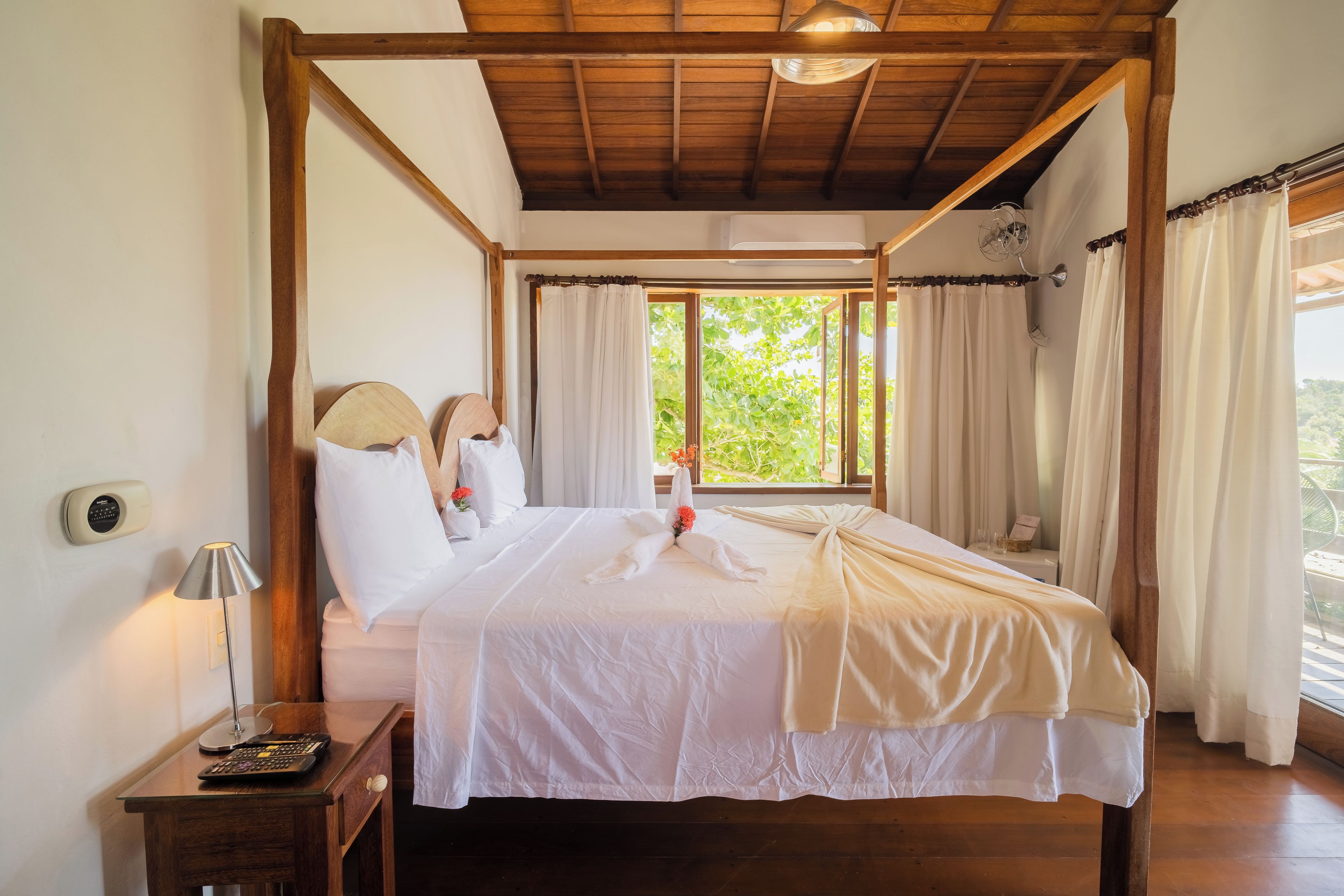 A four-poster bed with white linens, a bedside table with a lamp, and a view of greenery outside.