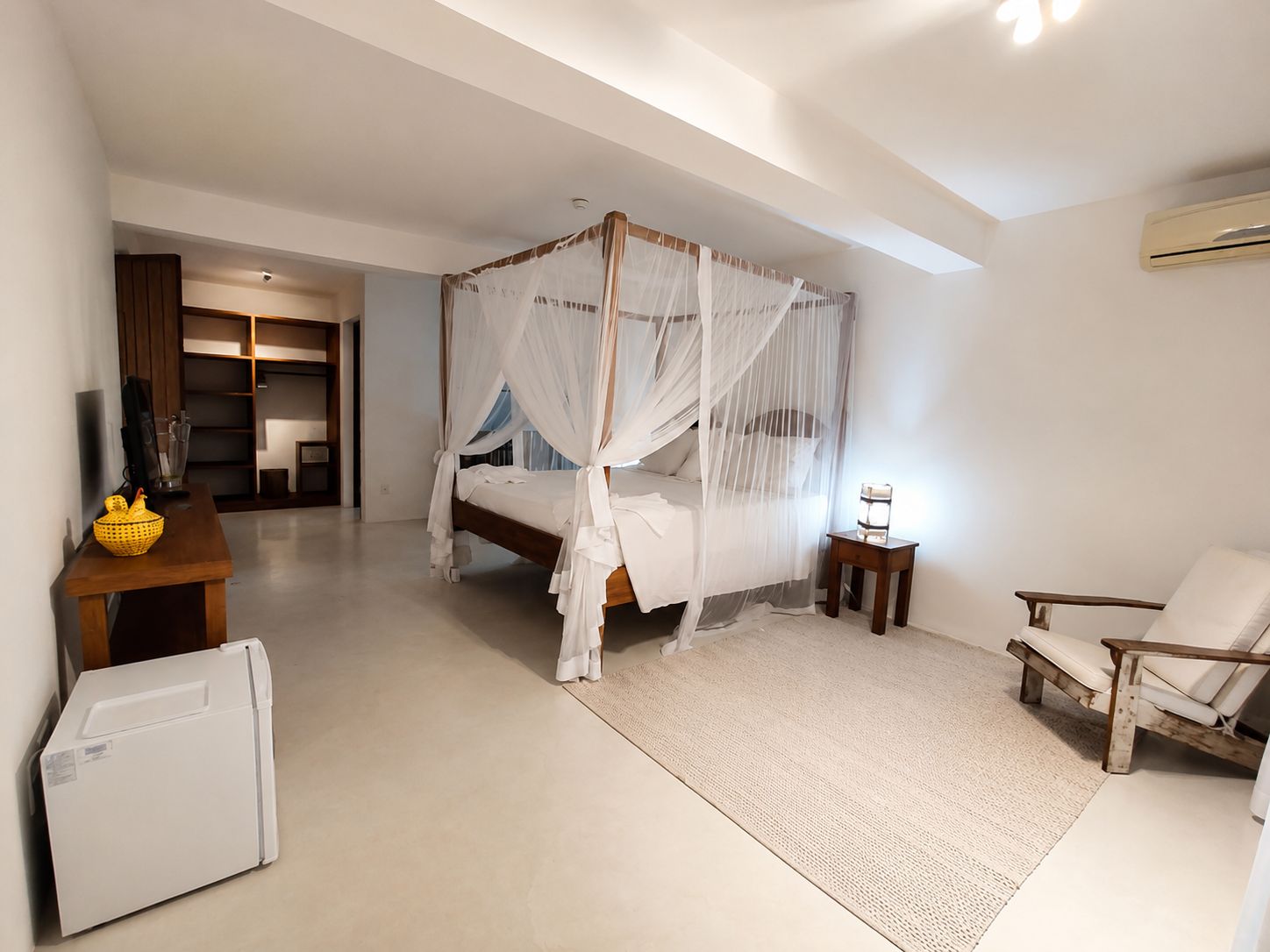 A bedroom with a four-poster bed, a small bedside table, a chair, a desk, and a refrigerator.