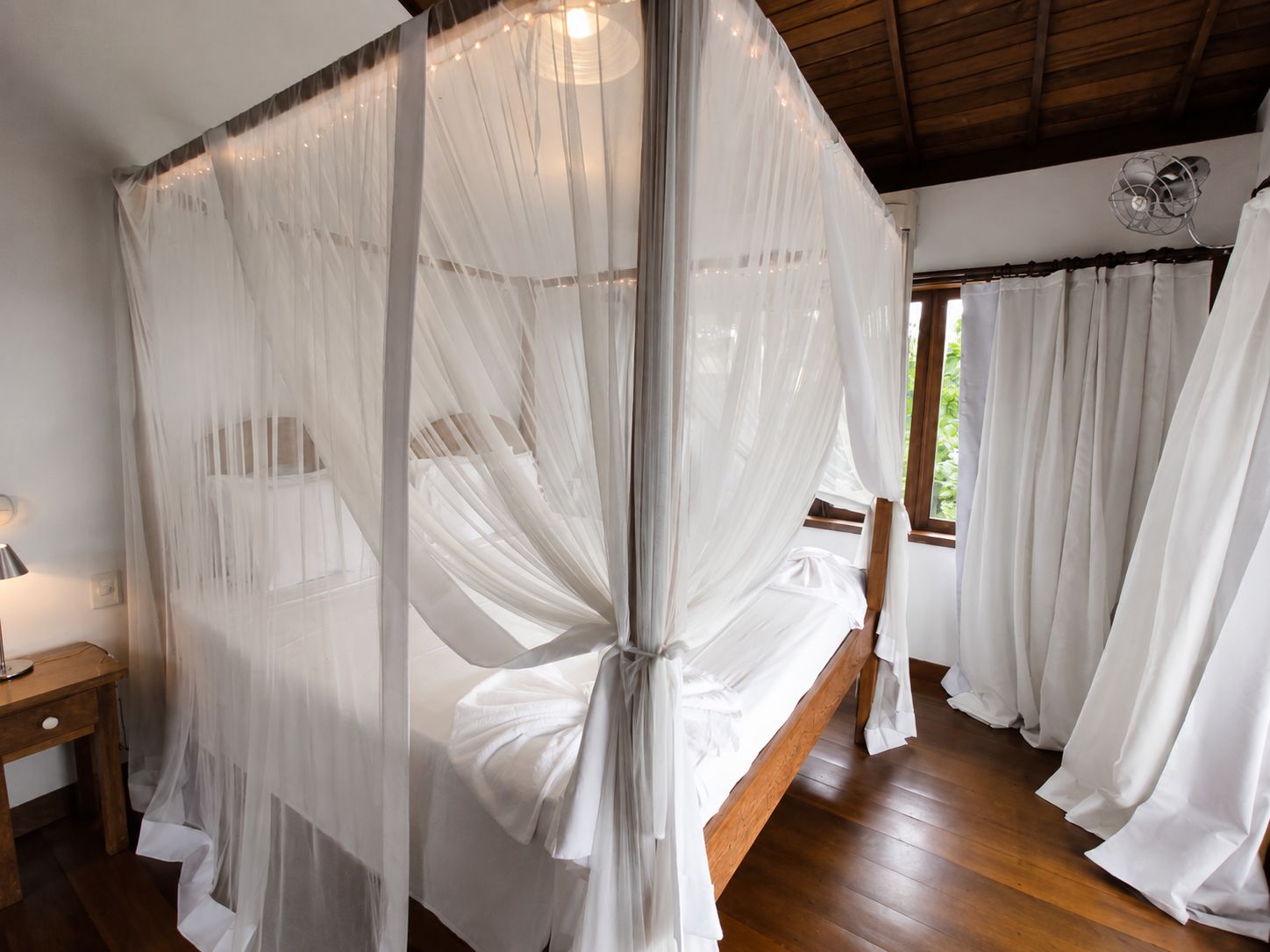 A room with a bed covered by a sheer canopy, wooden flooring, a bedside table, and curtains.