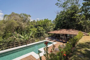 Piscina com deck de madeira, cercada por vegetação exuberante e um pequeno edifício com telhado em cerâmica.