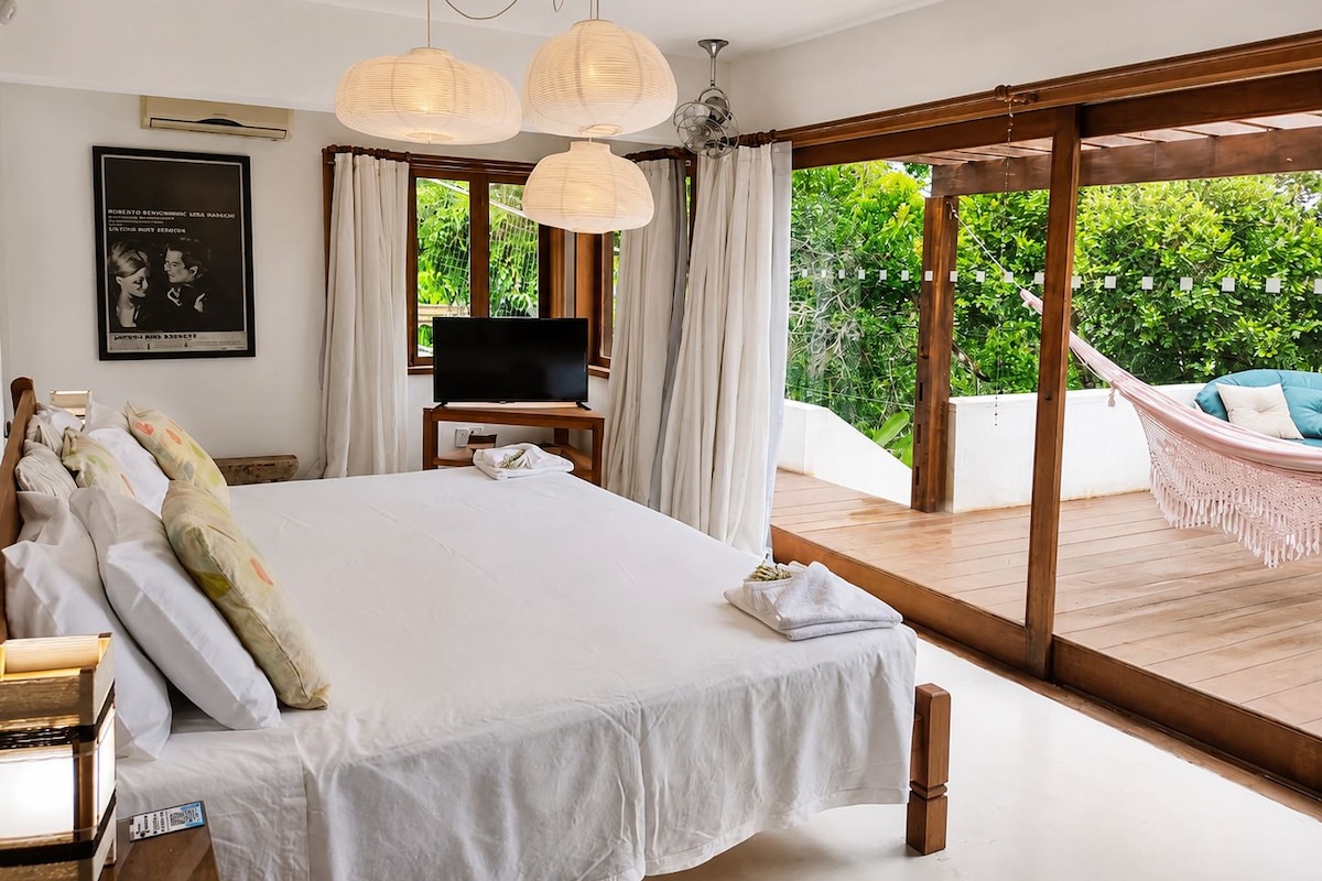 Un dormitorio con una cama grande, un televisor sobre un mueble y vistas a un jardín exuberante a través de una puerta corrediza de vidrio.