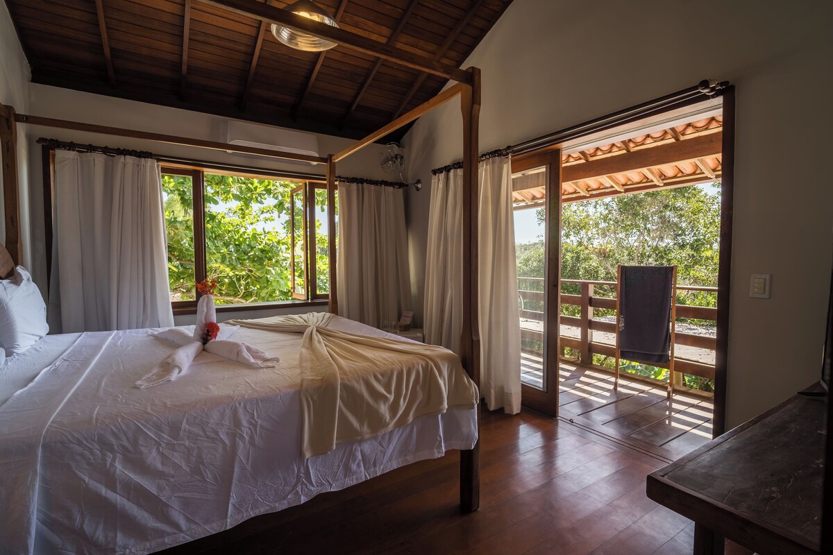Quarto de hotel com uma cama grande, cortinas, teto de madeira e vista para área externa através de uma porta de vidro.