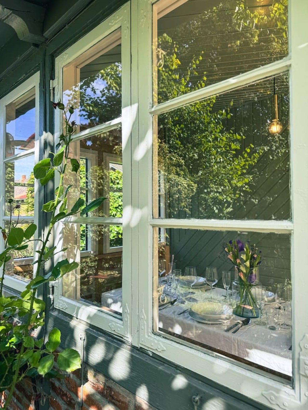 A room with large windows, a dining table set with glassware, and a hanging light fixture.