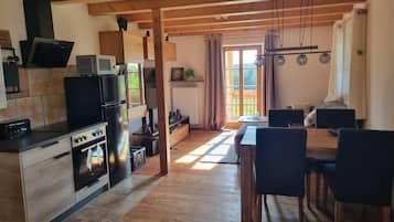 A modern kitchen with wooden beams, a dining area with a table and chairs, and a view through a window to the outside.