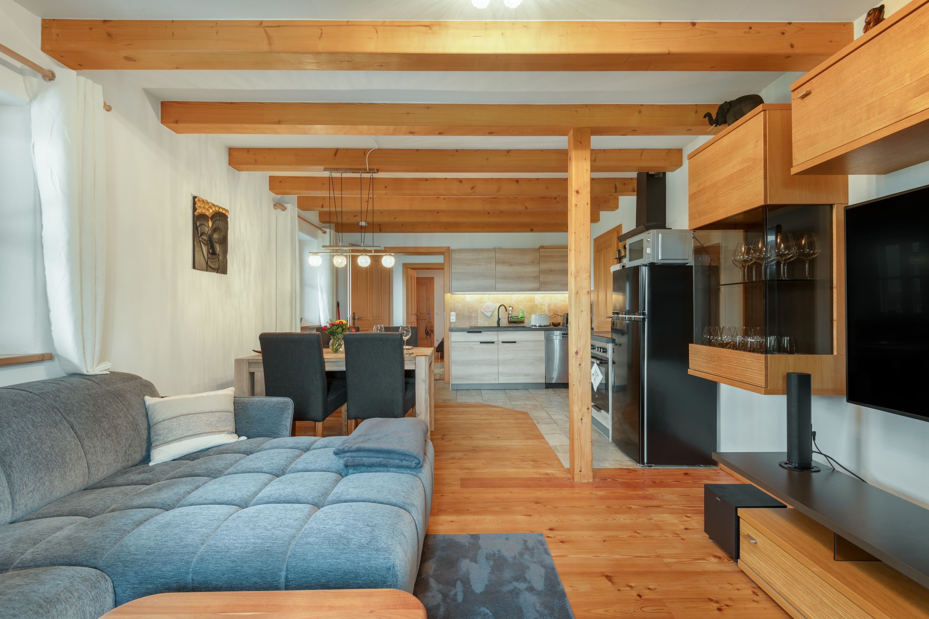 A modern living room with a grey sofa, wooden beams, a dining area with chairs, and a kitchen with a black refrigerator.