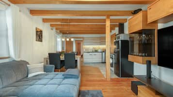 A modern living room with a grey sofa, wooden beams, a dining area with chairs, and a kitchen with a black refrigerator.