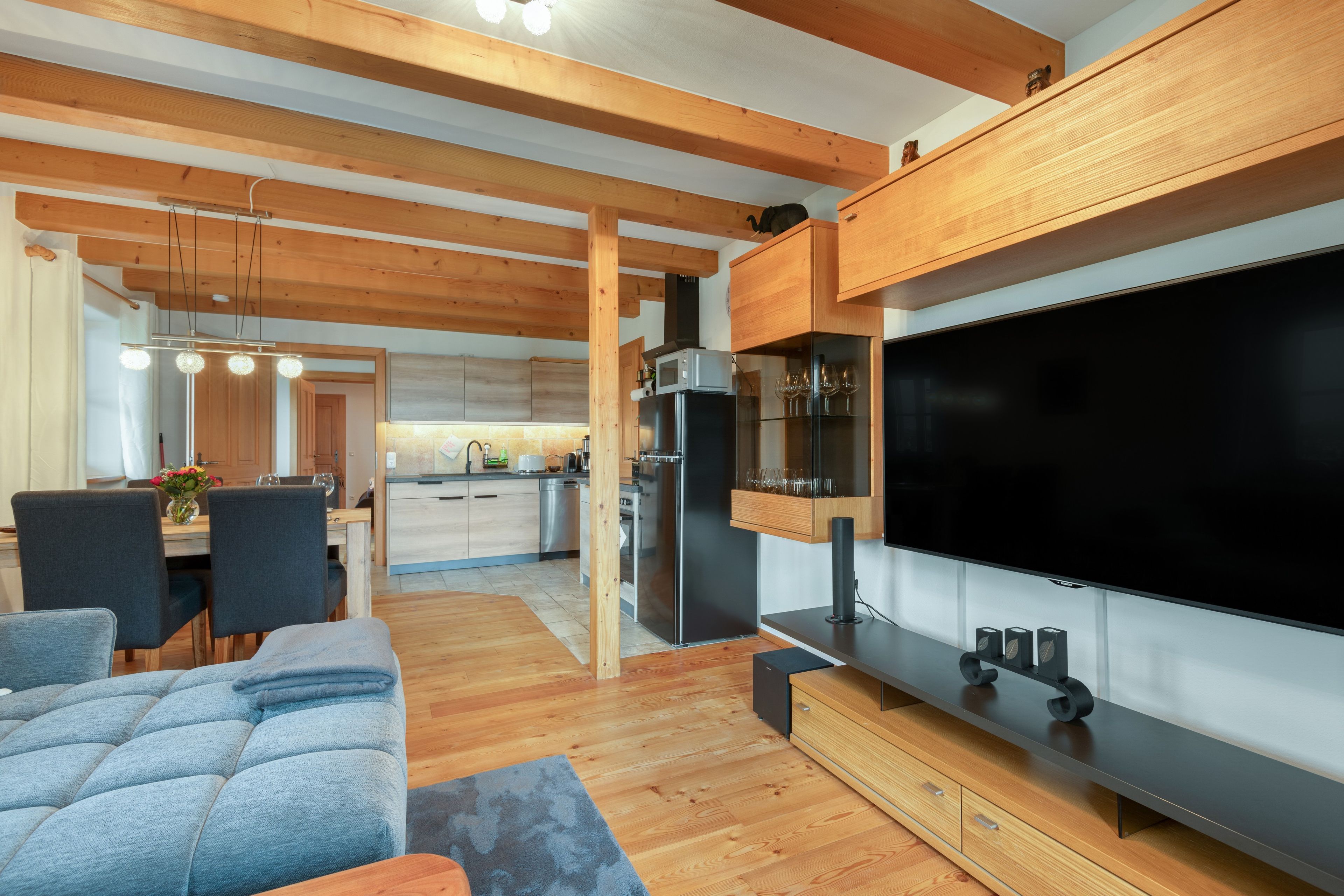 Ein modernes Wohnzimmer mit Flachbildfernseher, Holzbalken, einer Essecke und einer Küche im Hintergrund.