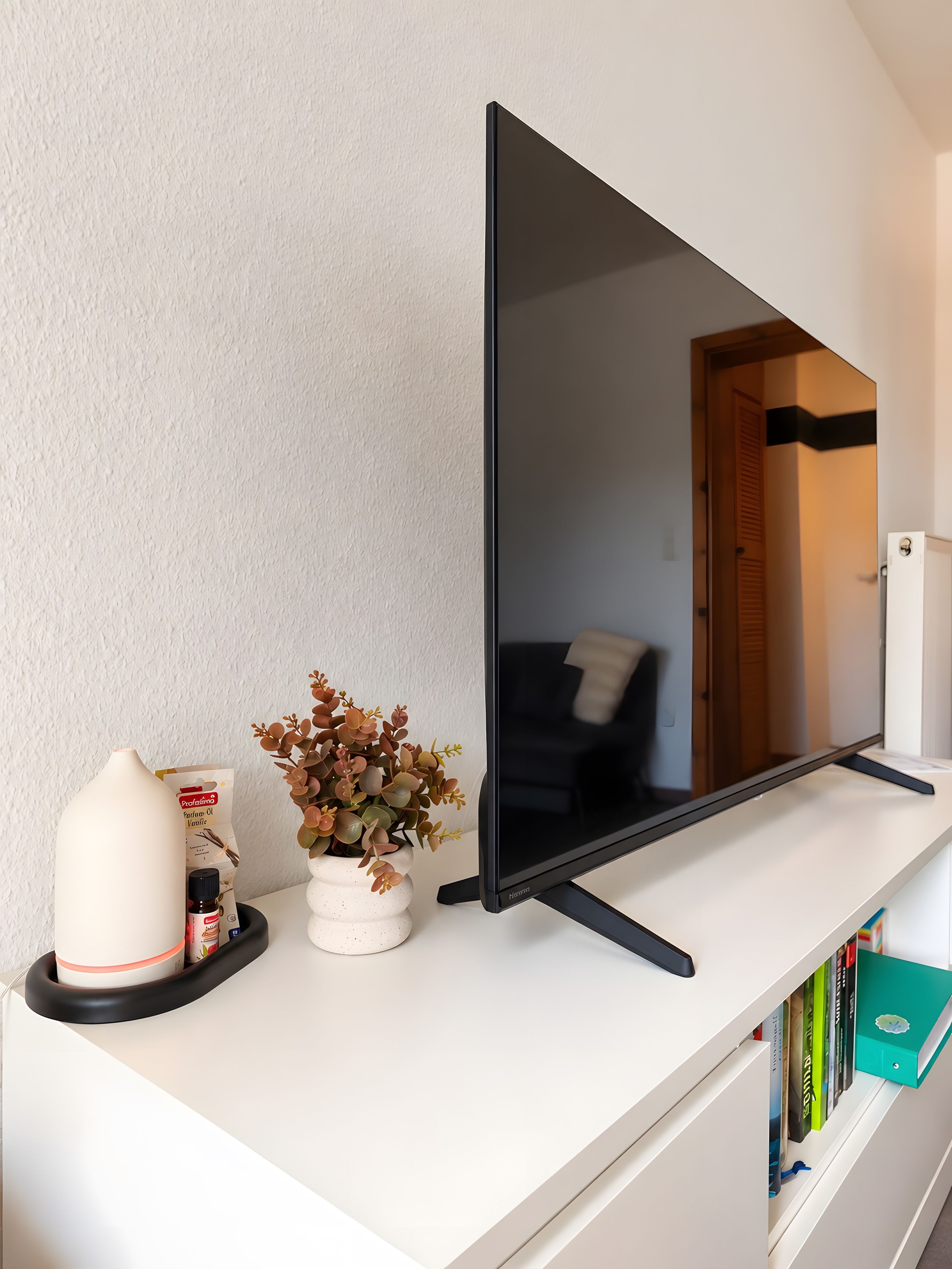 Ein Flachbildfernseher auf einem weißen Regal, eine weiße Vase mit getrockneten Blumen, ein weißer Behälter mit Etikett und ein Buch.