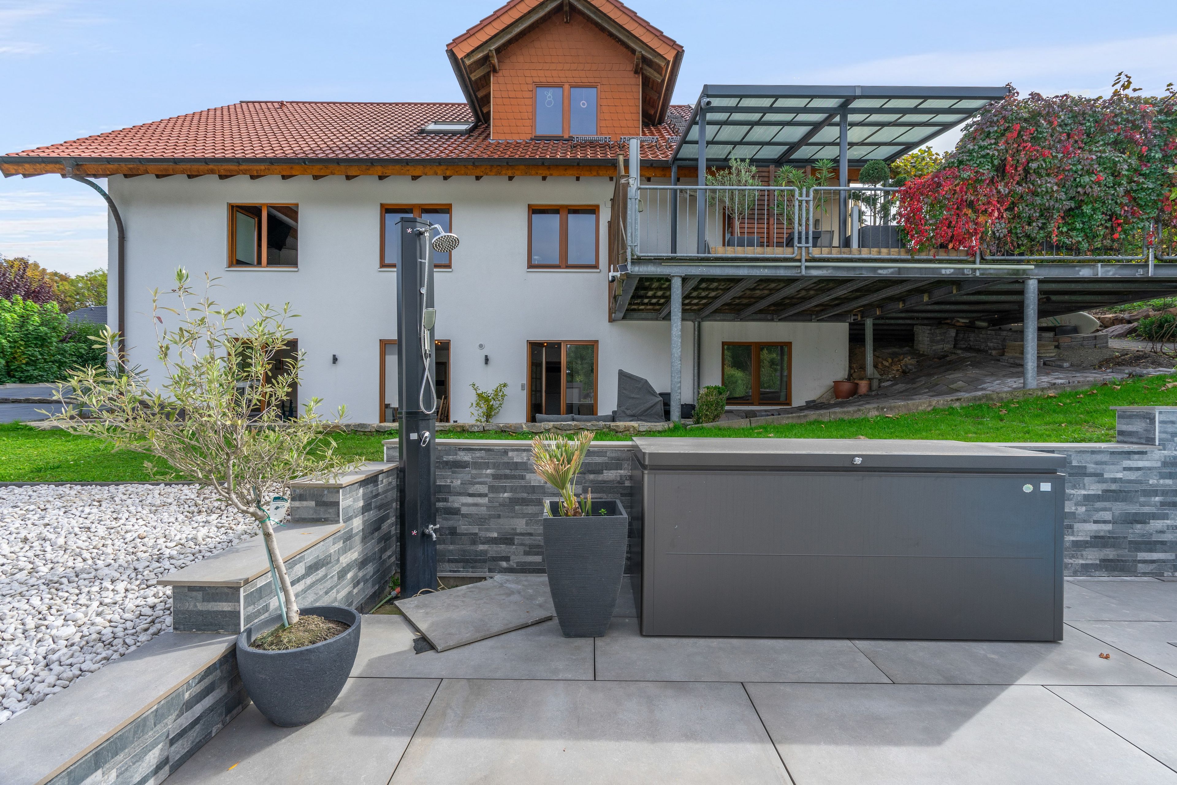 Ein modernes Haus mit überdachter Terrasse, einer großen grauen Garagentür und einem kleinen Baum in einem Blumentopf.