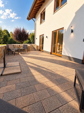 A patio area with stone flooring, outdoor furniture, and a view of a modern building with large windows and a stone wall.