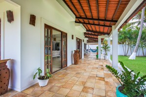 Área externa coberta com piso de azulejos, plantas em vasos e vista para o verde ao redor.