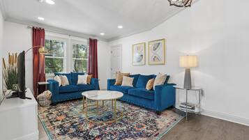 Un salon avec deux canapés bleus, une table basse blanche, un tapis à motifs, un lampadaire et deux œuvres d’art encadrées accrochées au mur.