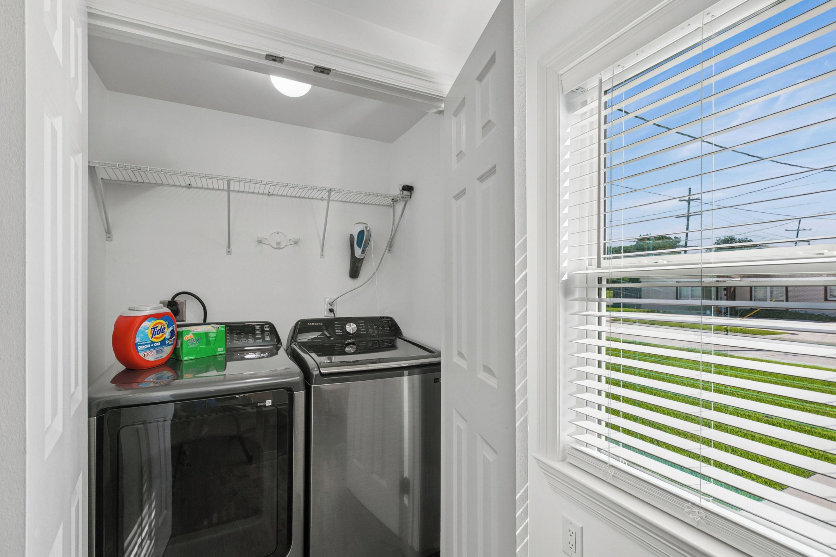 A laundry area with a washing machine and dryer, a drying rack, and a window with blinds.