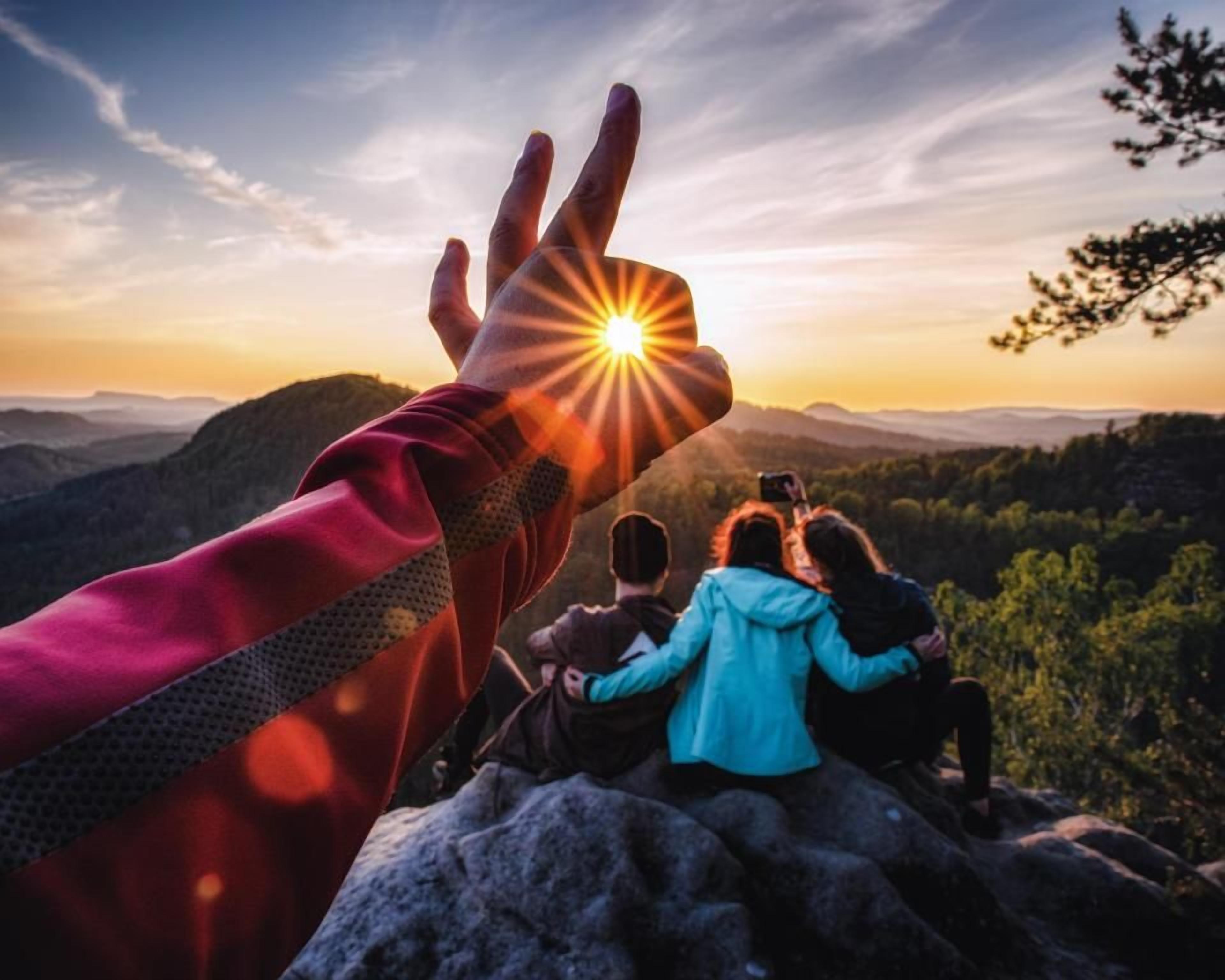 Una mano con un sol de fondo, un grupo de personas sentadas sobre una roca y un paisaje montañoso.