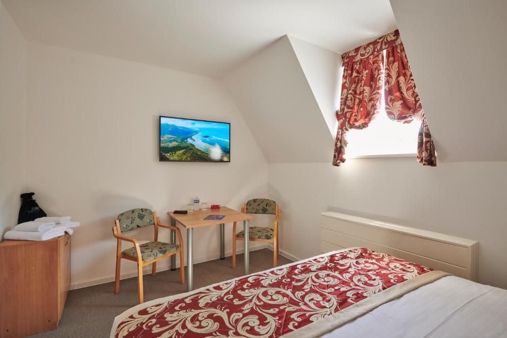 Ein Schlafzimmer mit einem Bett, einem Schreibtisch, zwei Stühlen, einem fenster mit Vorhängen, einem Bild an der Wand und einem kleinen Schrank.