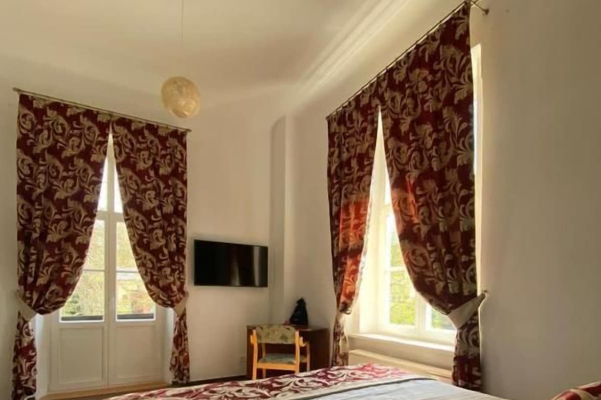 Quarto de hotel com uma cama grande, duas janelas com cortinas, uma TV de tela plana e uma mesa pequena.