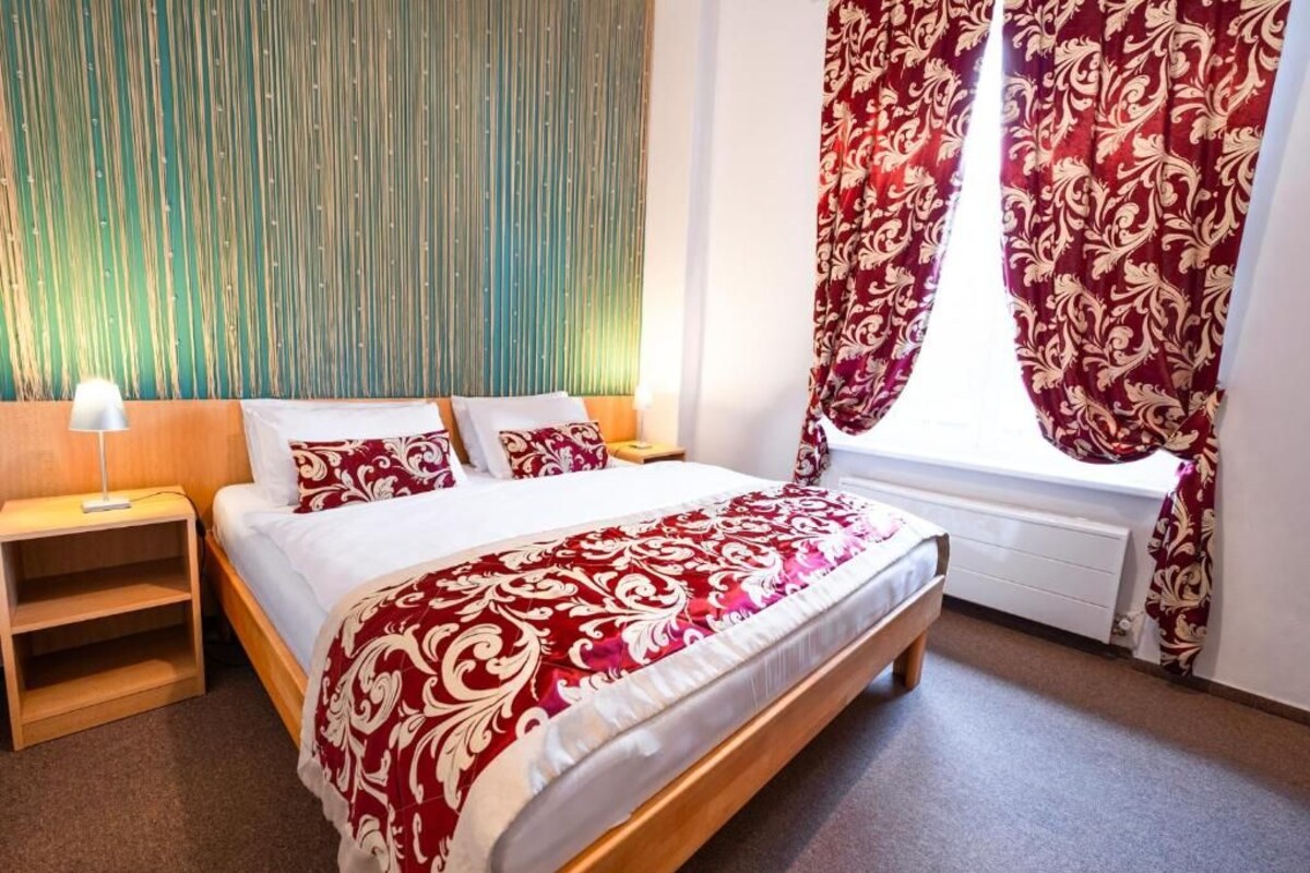 Quarto com cama, mesa de cabeceira, janela com cortinas e parede com linhas verticais.