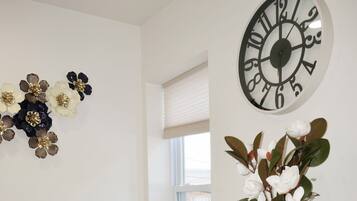 A modern living room with a sofa, a decorative wall clock, a vase with flowers, and a window with blinds.